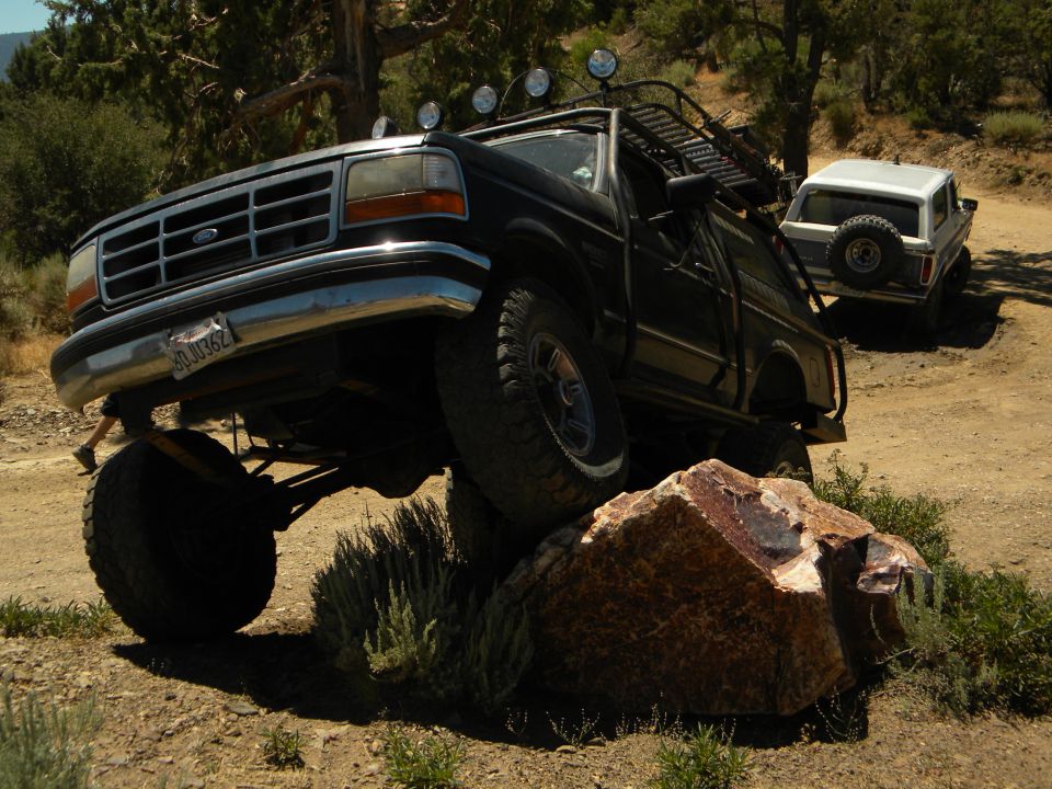 Black Ford truck with lifted suspension driving over a rock, another white SUV in background