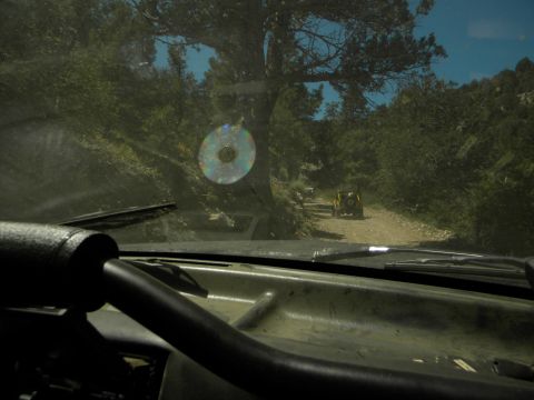 1992 Ford Bronco - A yellow SUV drives down a dusty road surrounded by trees, with another vehicle visible in the distance.
