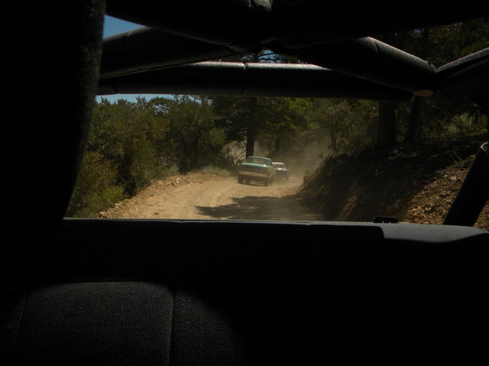 Two trucks drive on a dusty dirt road through trees. One truck is green
