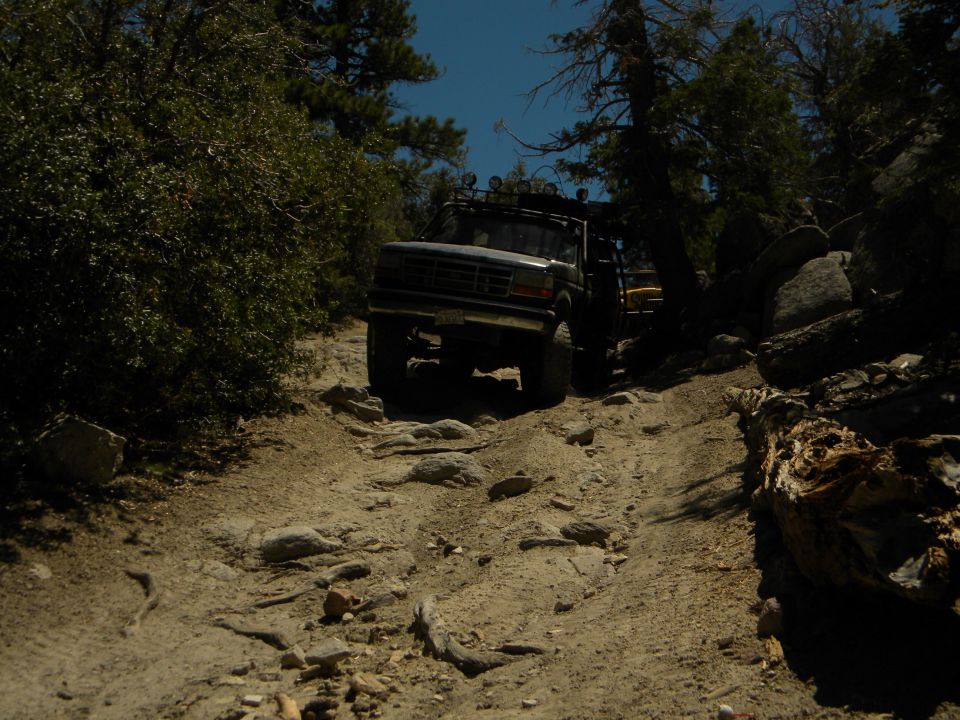 Truck climbing a rocky trail in the woods. Another vehicle is visible behind it