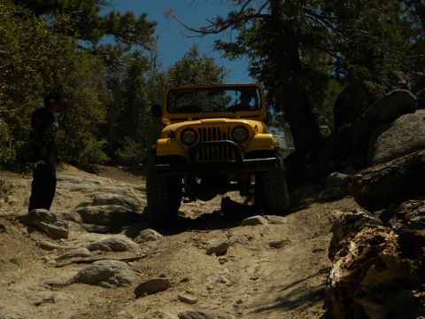 1992 Ford Bronco - A yellow Jeep CJ-7 navigates a challenging, rocky off-road path under a clear blue sky.