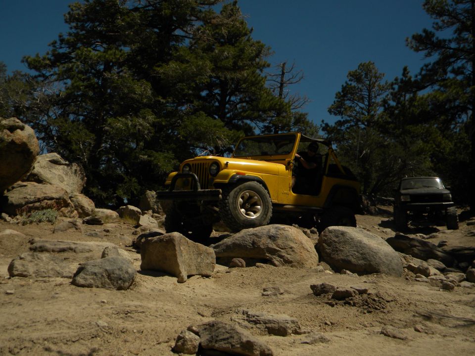 Yellow Jeep Wrangler on rocky terrain, driver visible, black SUV in background