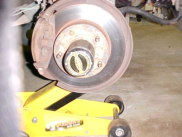 Close-up of a vehicle's front hub and brake rotor assembly, supported by a yellow jack. The hub has a yellow and black selector