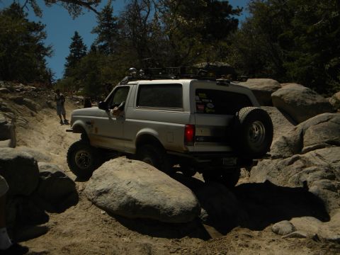 1992 Ford Bronco - A white 1992 Ford Bronco is shown off-roading on a rocky, dirt path with trees and boulders in the background.