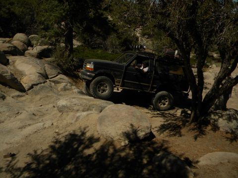1992 Ford Bronco - A black Ford Bronco is shown off-roading on a rocky terrain, with trees and boulders in the background.