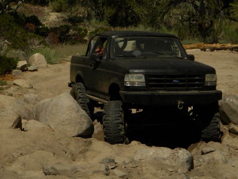 1992 Ford Bronco - A black 1992 Ford Bronco is shown off-roading on a dirt path with large rocks.