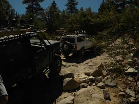 1992 Ford Bronco - Two SUVs navigate a challenging, rocky trail through a wooded area on a sunny day.