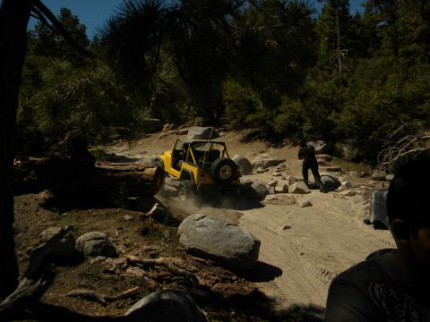 1992 Ford Bronco - A yellow Jeep Wrangler navigates a rocky off-road trail surrounded by trees and boulders.