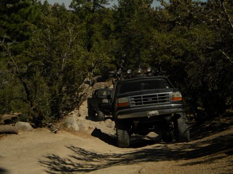 1992 Ford Bronco - Two off-road vehicles, including a 1992 Ford Bronco, navigate a rocky, wooded trail on a sunny day.
