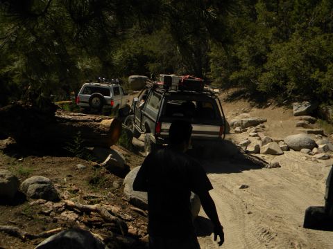 1992 Ford Bronco - Two off-road vehicles navigate a challenging, rocky path through a wooded area.