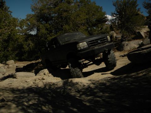 1992 Ford Bronco - A black 1992 Ford Bronco navigates a challenging off-road trail with large rocks and trees.