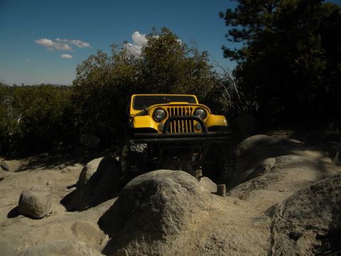 1992 Ford Bronco - Yellow Jeep CJ off-roading on a rocky trail under a blue sky with clouds.