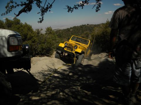 1992 Ford Bronco - A yellow Jeep Wrangler tackles a challenging rocky trail, with a white truck partially visible in the foreground.