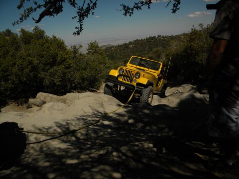 1992 Ford Bronco - A yellow Jeep Wrangler navigates a challenging rocky trail under a clear blue sky.