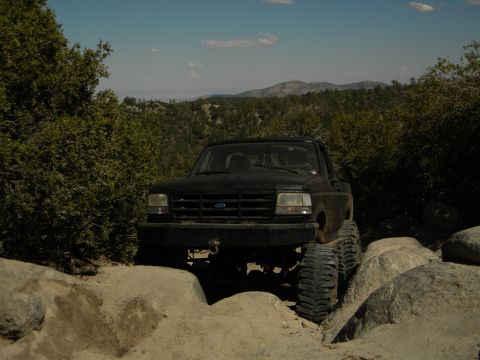 1992 Ford Bronco - A black Ford pickup truck is shown off-roading on a rocky trail with hills in the background.