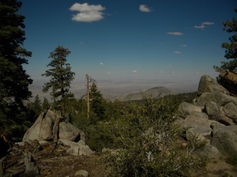 Mountain landscape with trees, rocks, and a distant valley under a blue sky with clouds. Watermark reads www.s