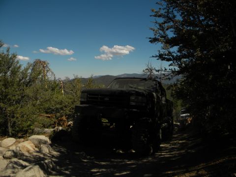 1992 Ford Bronco driving on a rocky dirt road with trees and mountains in the background. Another vehicle is v