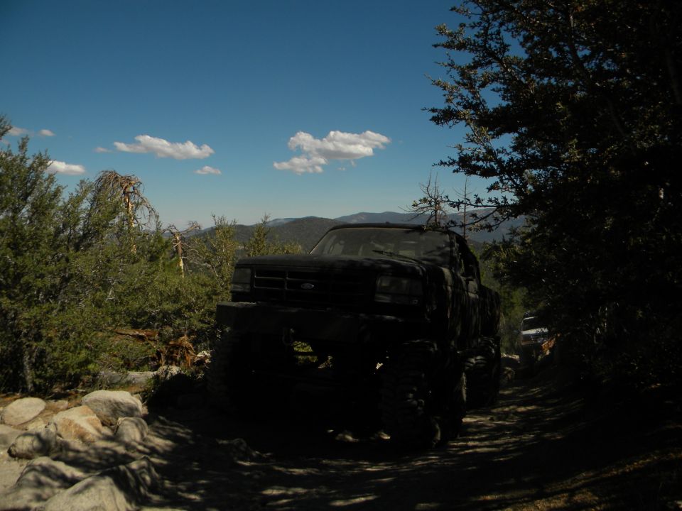 1992 Ford Bronco driving on a rocky dirt road with trees and mountains in the background. Another vehicle is visible ahead