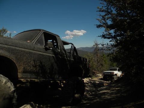 Muddy off-road truck in foreground, white SUV on trail behind it, trees and mountains in background