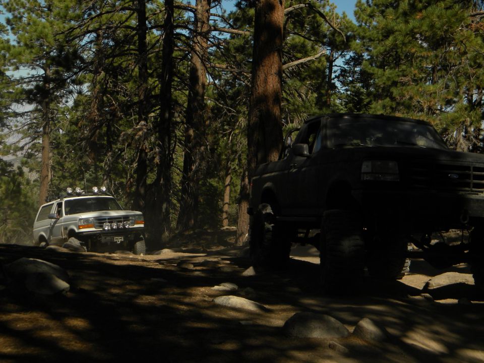 White 1992 Ford Bronco with roof lights drives on a dirt path in a forest, with a dark truck in the foreground
