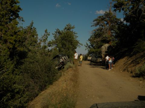Off-road vehicle on its side in bushes next to a dirt road with people standing nearby