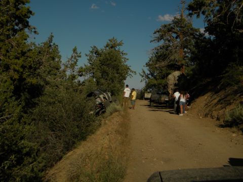Off-road vehicle tipped on its side in bushes next to a dirt road with people standing nearby