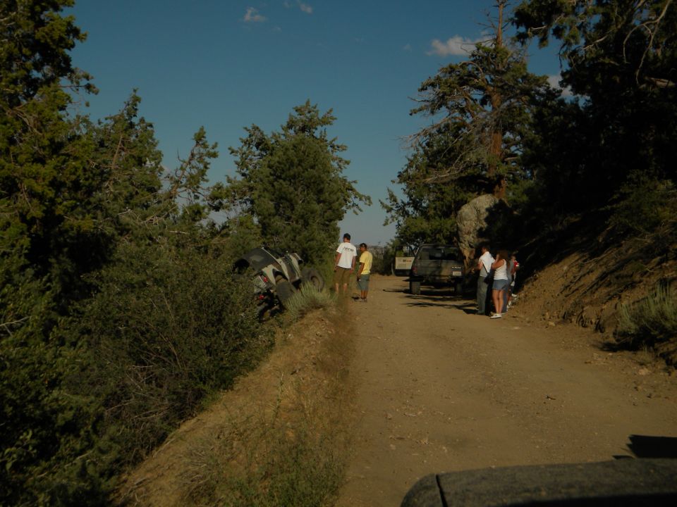 Off-road scene with a vehicle on its side in bushes and another vehicle parked on a dirt road with people standing around