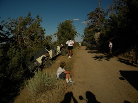 Camouflaged off-road vehicle tilted on a dirt embankment, with people standing on the road