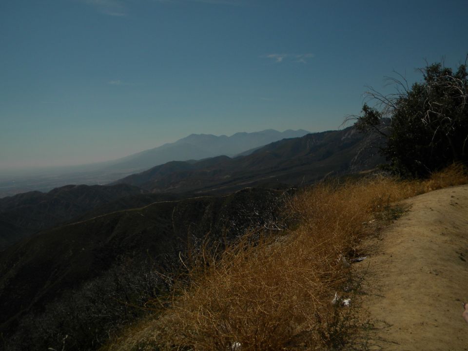 Mountain landscape with dry grass in the foreground and distant mountains under a clear blue sky. Watermark reads SUPERMOTORS