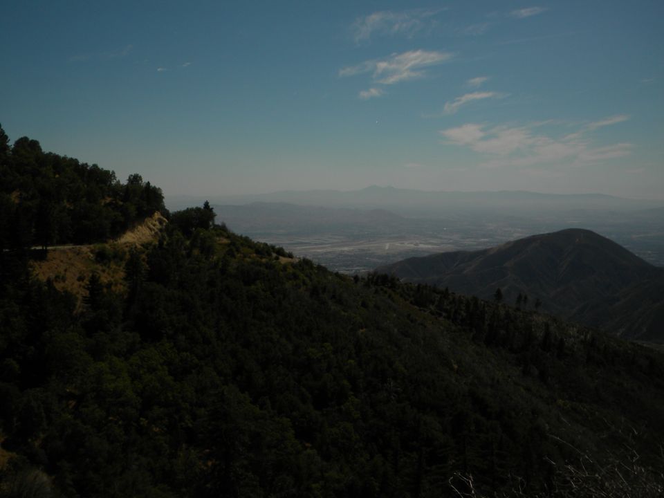 Mountainous landscape with a valley and distant haze under a blue sky with clouds. Watermark at bottom right