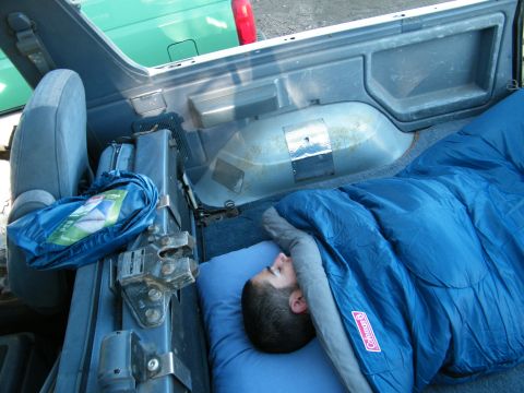 1982 Ford Bronco - A person sleeps in a blue sleeping bag inside the cargo area of a 1982 Ford Bronco.