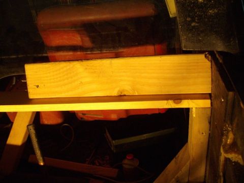 1978 Ford Bronco - Dimly lit shop shot of a windshield frame or glass panel being test-fit against a wooden jig built from fresh 2x4 lumber, with a red toolbo…