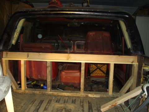 1978 Ford Bronco - Rear view into a gutted black Ford Bronco with the top removed, showing a rough-framed lumber bulkhead built across the cab where the rear…