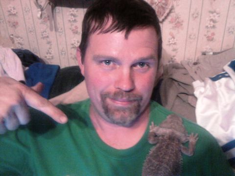 1989 Ford Bronco - Indoor selfie of a person in a green T-shirt with a goatee, pointing at a bearded dragon lizard perched on their shoulder, in front of flor…