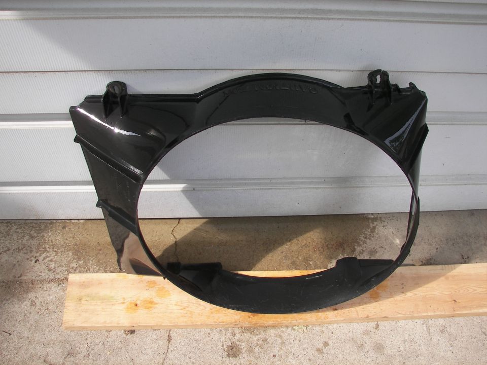 Black plastic radiator fan shroud with mounting tabs, resting on wood planks