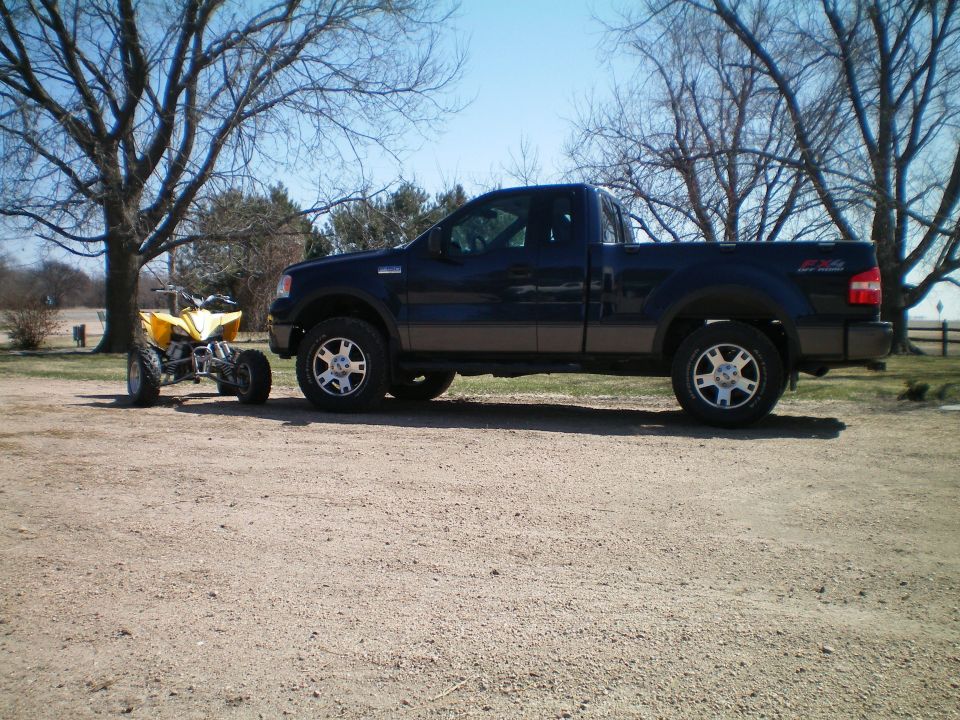 Dark blue Ford F-150 pickup truck, side view, parked on gravel next to a yellow ATV