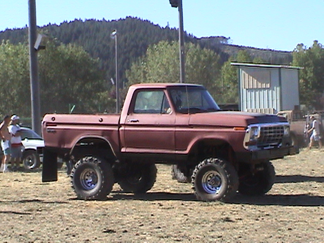 Lifted maroon vintage Ford pickup on oversized mud tires at a dirt event grounds