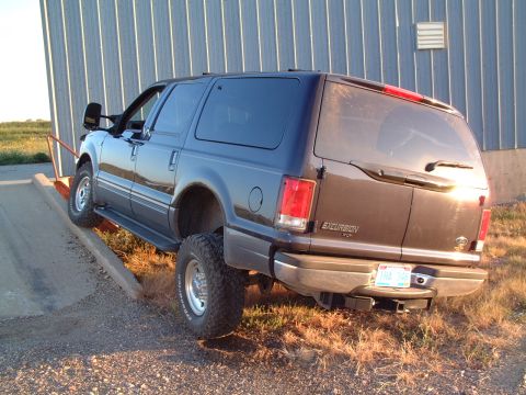 1986 Ford Bronco - A lifted Ford Excursion is positioned on a ramp, partially obscuring the lower portion of a corrugated metal building.