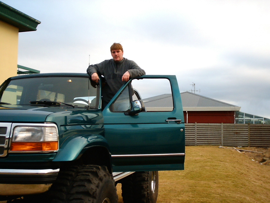 Teal lifted Ford Bronco with door open, man leaning on frame, big off-road tires, parked on grass.