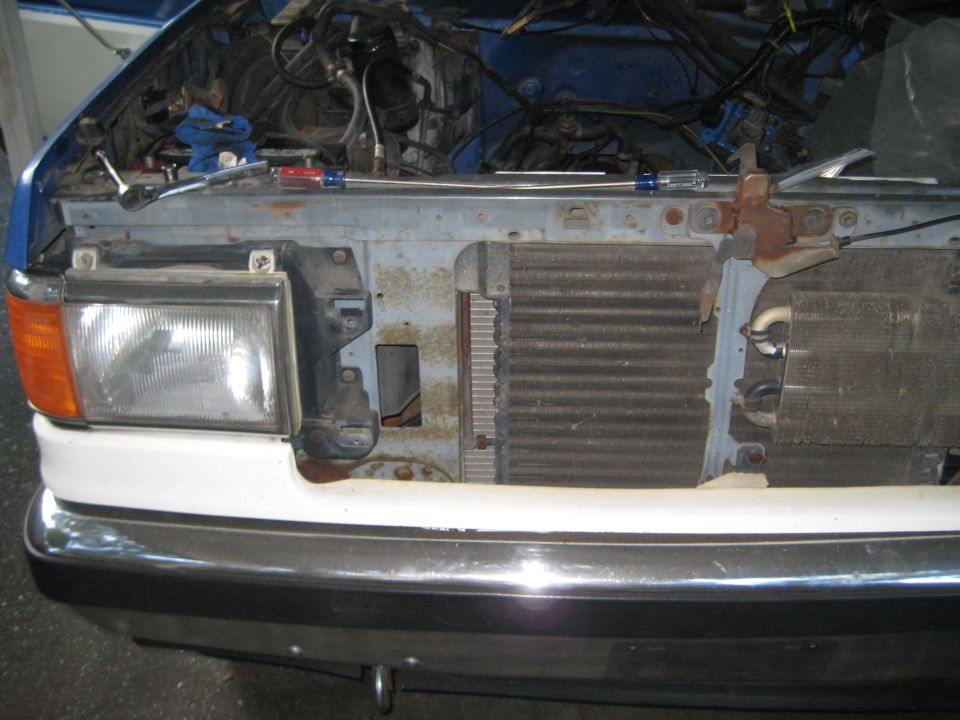 Front end of a blue and white vehicle with hood open, showing exposed engine and radiator. Tools are on the engine bay