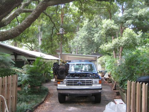 1988 Ford Bronco - A blue Ford Bronco sits in a driveway with its hood open, while an engine is suspended from a tree branch overhead.
