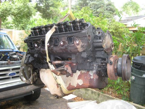 1988 Ford Bronco - Engine removed from a vehicle, suspended by straps. A blue truck is partially visible in the background.