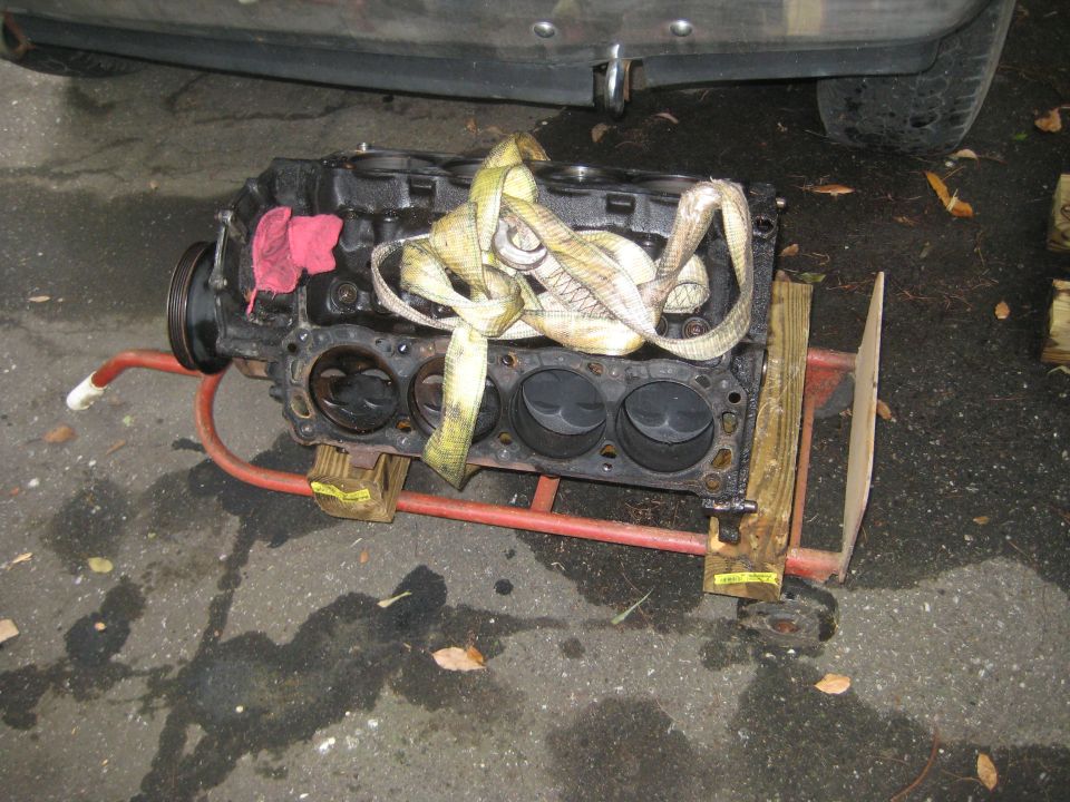 Engine block on a red dolly, secured with straps and wood, with a red rag. Part of a vehicle is visible above