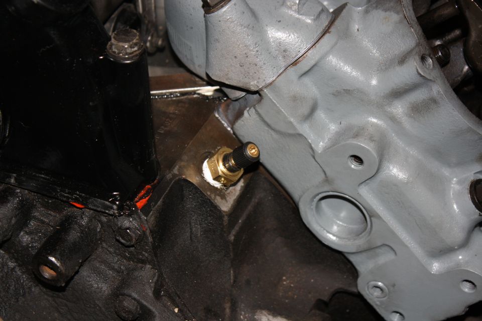 Close-up of a brass sensor screwed into a gray engine block, with black painted metal parts nearby