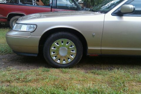 2000 Mercury Grand Marquis - A gold sedan is parked next to a red pickup truck, showing the front passenger side wheel and headlight.