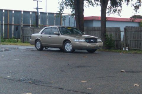 2000 Mercury Grand Marquis - Tan 2000 Mercury Grand Marquis parked outdoors in front of a fence and trees.