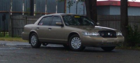 2000 Mercury Grand Marquis - A tan sedan is parked on a wet surface with a fence and trees in the background.