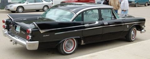 1991 Ford Crown Victoria - A classic black Dodge sedan with distinctive tail fins is parked in an outdoor lot, with other vehicles and pedestrians in the background.