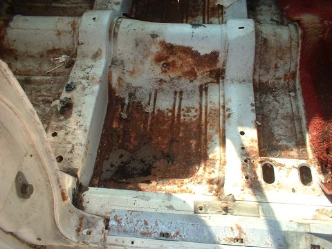 1967 Ford Galaxie - The interior floor pan shows significant rust and deterioration, with a glimpse of red carpet in the background.