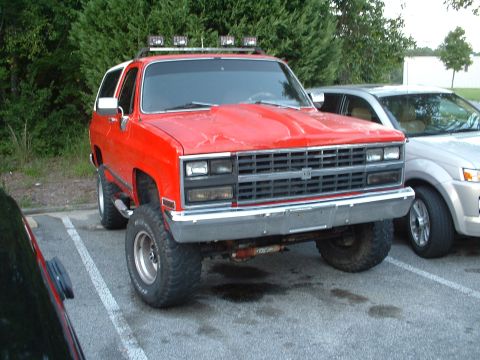 1994 GMC Suburban - This lifted red SUV features off-road lights mounted on a roof rack, parked next to another vehicle.